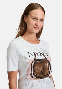 Hvid T-shirt med et grafisk print af en brun håndtaske med nitter, ledsaget af ordet "JOYFUL" i fed sort skrift. Korte ærmer.
