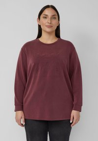 Kobieta z prostymi ciemnymi włosami, ubrana w bordową bluzkę z długim rękawem z subtelnym wytłaczanym wzorem, stojąca na tle jednolitego jasnoszarego tła.