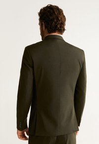 Giacca da suit verde oliva, vestibilità sartoriale, spacco singolo, revers a notched, texture del tessuto liscia, dettagli con tre bottoni sui polsini, vista posteriore.