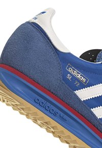 Niebiesko-białe buty sportowe Adidas SL '72 z zamszowym obcasem, czerwoną lamówką na niebieskiej śródpodeszwie oraz beżową, fakturowaną gumową podeszwą zewnętrzną.