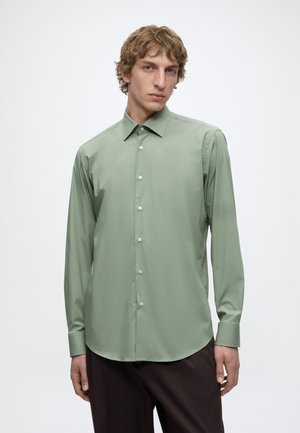 Jeune homme aux cheveux bouclés portant une chemise vert clair à manches longues boutonnée et un pantalon foncé, sur un fond uni.