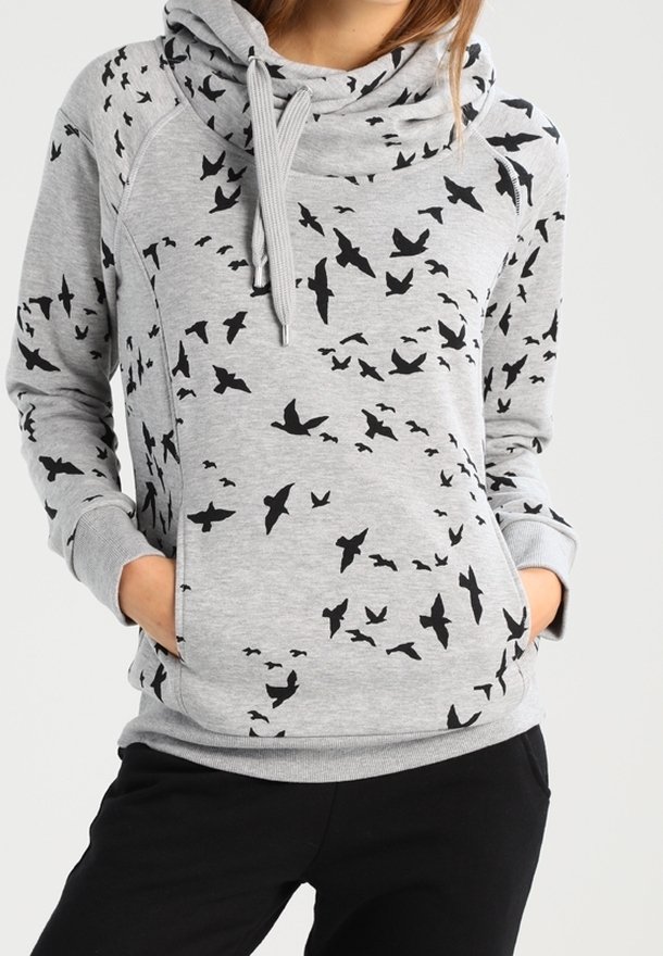 Personne portant un sweat à capuche gris clair avec des silhouettes d'oiseaux noirs et les mains dans les poches avant, associé à un pantalon noir.
