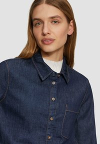 Granatowa koszula zapinana na guziki z dżinsu, z ostrym kołnierzykiem, kieszenią na piersi oraz brązowymi detalami stębnowania. Tkanina ma gładką teksturę.