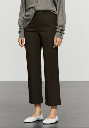 Personne portant un pantalon droit marron foncé, un haut gris à manches longues et des ballerines blanches, debout sur un sol gris.