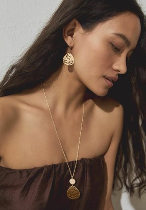 Gold jewelry includes a textured, teardrop pendant with engraved floral patterns and a small round accent. Appears worn on bare skin.