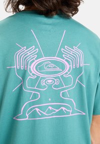 Tyrkysové bavlněné tričko s velkým růžovým grafickým motivem rukou a scenérií hor, umístěným uprostřed zad, s hladkou texturou.