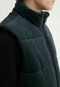 Dunkelgrüwestes Cord-Vest mit hohem Kragen, Reißverschluss und gesteppten Einsätzen. Strukturierter Stoff mit einer strukturierten Silhouette.