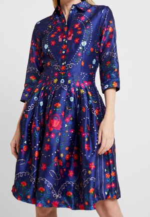 Woman wearing a knee-length, navy blue dress with red, pink, and yellow floral patterns and three-quarter sleeves, standing with one hand by her side.