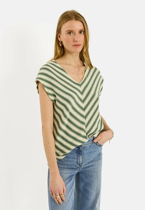 Femme portant un haut sans manches en maille rayé vert et crème avec un col en V et un jean bleu, debout avec une main dans la poche sur fond blanc.