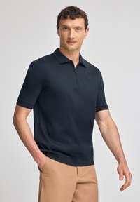 Mörkblå kortärmad pikétröja med dragkedja i kragen, tillverkad av ett mjukt tyg. Har en figurnära design och en diskret logotyp på bröstet.