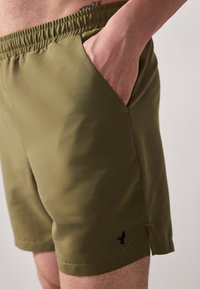 Olive green shorts with an elastic waistband, side pockets, and a small black logo. Smooth fabric with a slight sheen and a straight cut.