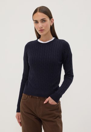 Joven mujer con cabello castaño liso, vestida con un suéter de punto grueso azul marino sobre una camisa blanca y pantalones marrones, de pie con una mano en el bolsillo.