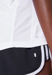 Valkoinen urheilupaita, jossa on verkkokangaspaneelit ja "AEROREADY" -logo. Mustat shortsit, joissa on valkoiset sivuraitat ja sileä tuntuma.