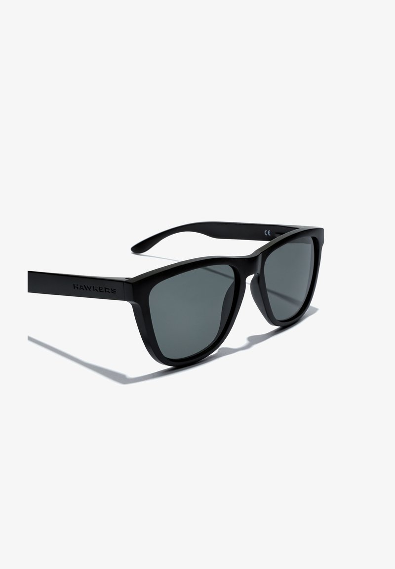 Črne plastične sončna očala s sivimi lečami, ki imajo klasično kvadratno zasnovo. Ročaji imajo mat površino, na eni strani pa je vtisnjen znak blagovne znamke "HAWKERS".
