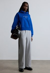 Modrá mikina s kapucňou a bielym logom, doplnená sivými širokými teplákmi; čierna taška a robustné čierne topánky dokončujú outfit.