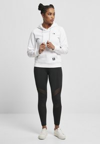 Hvit hettegenser med svart logo, kombinert med svarte leggings med gjennomsiktige paneler. Modellen står i hvite joggesko mot en nøytral bakgrunn.