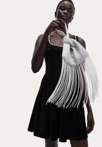 Černé sametové šaty s hranatým výstřihem, zdobené flitry. Drží stříbrný třásňový doplněk s hladkým povrchem.