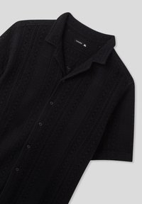 Camisetta nera a maniche corte con un design a maglia texture, caratterizzata da motivi a giorno, colletto e bottoni neri sul davanti.