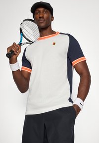 Ανδρικό κοντομάνικο μπλουζάκι τένις σε λευκό πλέγμα με ναυτικές λεπτομέρειες και πορτοκαλί περιθώριο, συνδυασμένο με μαύρα σορτς και μία ρακέτα τένις.
