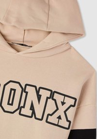 Felpa beige con grafica nera "BRONX". Materiale morbido con una finitura strutturata e una striscia nera a contrasto sulle maniche.