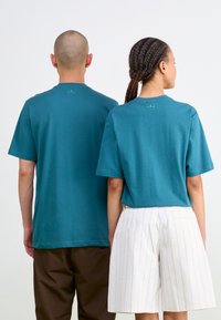Teal cotton t-shirts with short sleeves, back branding detail. One partner wearing brown shorts; the other in striped white shorts.