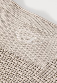 Lys beige strikket stof med et struktureret mønster, der har et lille, broderet logo. Udhalsningen er bred med ribbede detaljer.