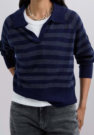 Henkilö, jolla on yllään tummansininen ja harmaa raidallinen pitkähihainen poolopaita valkoisen paidan päällä, harmaat farkut ja paksu hopeinen ketjukaulakoru.