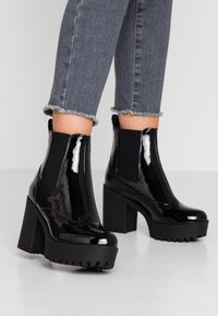 Botines de charol negros con tacones gruesos, paneles laterales elásticos y suelas de goma negra texturizada, combinados con jeans grises deshilachados.