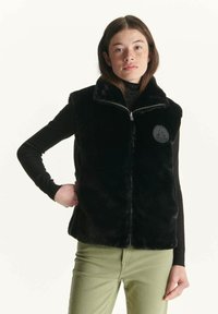 Chaleco de piel sintética negra con cremallera frontal, cuello alto y un logo circular. Llevado sobre un jersey de cuello alto negro y pantalones verdes. Textura suave.