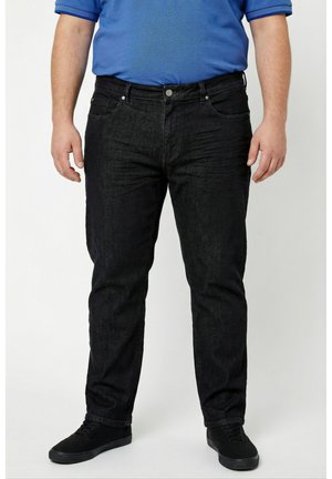 Mann trägt schwarze Slim-Fit-Jeans, schwarze Sneakers und ein blaues Poloshirt mit kurzen Ärmeln, steht vor einem einfarbigen Hintergrund.