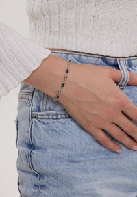 Srebrna bransoletka łańcuszkowa z zielonymi i niebieskimi kamieniami szlachetnymi, z przeplatającymi się owalnymi ogniwami, noszona na nadgarstku, zestawiona z jasnoniebieskimi dżinsami.