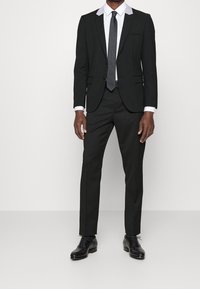 Zwart pak met een wit shirt met grijze accenten op de kraag, gecombineerd met een zwarte stropdas en gepolijste zwarte leren schoenen.