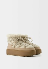Kremfarvede støvler med tekstureret pels, chunky beige platform og sorte og hvide snørebånd med mønster, samt gyldne metalaccenter.