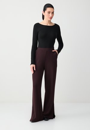 Vrouw staand met hand in de zak, gekleed in een zwart langermig shirt en wijde, hooggetailleerde donkergroene broek, tegen een effen achtergrond.