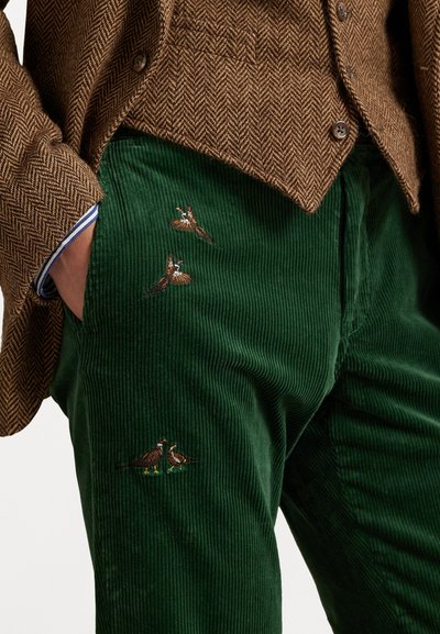 Green corduroy trousers featuring embroidered birds in brown, red, and green. Paired with a brown herringbone knit jacket.