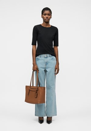 Woman wearing a black elbow-sleeve top and light blue wide-leg jeans, holding a brown leather tote bag, standing against a white background.