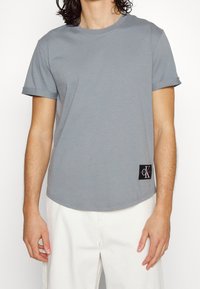 Hombre vestido con una camiseta de manga corta gris claro con un parche negro del logo de CK en el dobladillo, combinada con pantalones blanquecinos, de pie contra un fondo blanco.