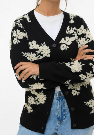 Femme les bras croisés portant un cardigan noir à motifs floraux blancs, une chemise blanche et un jean bleu.