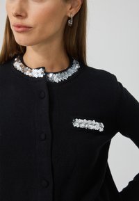 Czarny kardigan z kołnierzem i kieszenią ozdobionym cekinami. Posiada okrągły dekolt i zapięcie na guziki, wykonany z teksturowanej dzianiny.