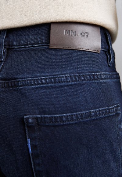 Tummat farkut, joissa on nahkainen etiketti, jossa lukee "NN. 07", näkyvä ommel ja huomaamaton taskutieto, esittelevät sileää ja rakenteellista tekstuuria.