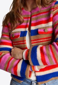 Femme portant une veste en laine rayée colorée avec des boutons dorés, bras croisés, montrant des rayures horizontales rouges, roses, bleues, beiges et orange.