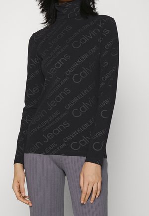 Woman wearing a black turtleneck covered with gray "Calvin Klein Jeans" text and gray ribbed pants, standing against a plain background.