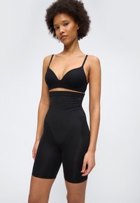 Γυναίκα με σγουρά μαλλιά που φοράει μαύρα σορτς σχήματος υψηλής μέσης και μαύρο σουτιέν, στέκεται μπροστά σε απλό ανοιχτόχρωμο φόντο.