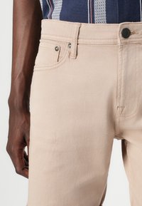 Pantalons en coton beige avec une texture lisse, dotés d'une fermeture à bouton, de poches latérales et d'une coupe ajustée à la taille.