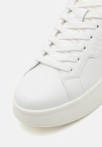 Hvit sneaker med glatt syntetisk overdel, rund tå og flate hvite snøringer. Har en teksturert såle og preget merkevaredetalj.