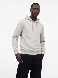 Homme portant un sweat à capuche gris clair avec cordons et un pantalon noir, debout avec une main dans la poche devant un fond blanc uni.