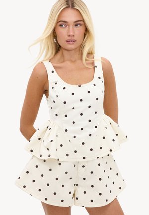 Femme aux longs cheveux blonds portant un top blanc sans manches à pois avec basque et un short assorti, regardant sur le côté.