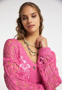 Cárdigan de punto rosa con patrón calado, que presenta un escote en V, cierre de botón y mangas largas. Múltiples collares de cuentas coloridas adornan el cuello.