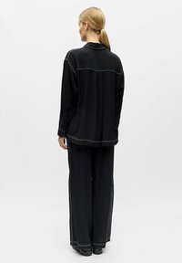 Femme aux cheveux blonds attachés en queue de cheval, vue de dos, portant un haut noir ample et un pantalon large noir avec des coutures blanches.