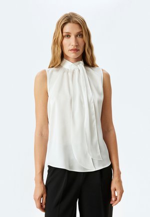 Weiße ärmellose Bluse mit hohem Kragen und Schleifendetail. Aus weichem, transparentem Stoff gefertigt. Taillierte Passform mit leichtem Fall, kombiniert mit dunklen Hosen.